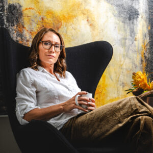 Subject poses seated in a wingback chair, before a colorful painting, holding a coffee.